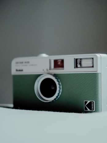 A vintage-style camera with a green leather texture on a neutral background. The camera has a distinctive circular lens and a small viewfinder, with branding on the front panel.