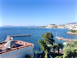 A coastal view with a pier extending into the sea, lined with umbrellas and sunbeds. In the foreground, there are palm trees and a building with a geometric roof. Across the water, a town with white buildings is nestled on a hillside, with mountains in the background under a clear blue sky.