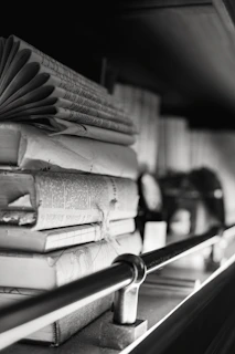 A stack of old, worn books is resting on a shelf. The books are in varying sizes and are stacked horizontally and vertically. The image is in black and white, adding a classic and timeless feel. There is a metal rod running horizontally in front of the books, which appears to be part of the shelf's design.