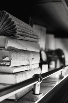 A stack of old, worn books is resting on a shelf. The books are in varying sizes and are stacked horizontally and vertically. The image is in black and white, adding a classic and timeless feel. There is a metal rod running horizontally in front of the books, which appears to be part of the shelf's design.