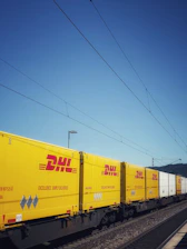 Yellow and white DHL freight containers are lined up on a train track under clear blue skies. Overhead electrical wires stretch across the scene, indicating a railway setting.