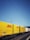 Yellow and white DHL freight containers are lined up on a train track under clear blue skies. Overhead electrical wires stretch across the scene, indicating a railway setting.