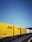 Yellow and white DHL freight containers are lined up on a train track under clear blue skies. Overhead electrical wires stretch across the scene, indicating a railway setting.