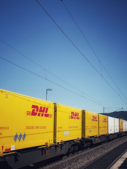 Yellow and white DHL freight containers are lined up on a train track under clear blue skies. Overhead electrical wires stretch across the scene, indicating a railway setting.
