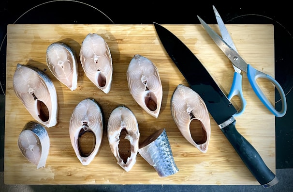 Several raw fish steaks are arranged on a wooden cutting board alongside a large kitchen knife and a pair of kitchen scissors with blue handles.