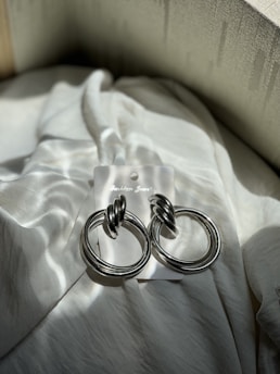 A pair of shiny, silver hoop earrings with intertwined design are placed on a soft, textured fabric. The earrings are mounted on a white card, likely the packaging, with light shadows cast upon them, creating a subtle interplay of light and texture.