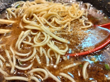 A close-up of a bowl of ramen noodles in a rich, dark broth. The noodles are tangled and partially submerged. A red spoon is visible on the right side, resting in the broth, and specks of seasoning are scattered throughout.