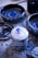 A stack of ceramic lids with colorful paint splatters rests on a paint-speckled surface. Ceramic bowls with intricate blue designs are placed around, suggesting an artist's workspace.