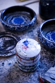 A stack of ceramic lids with colorful paint splatters rests on a paint-speckled surface. Ceramic bowls with intricate blue designs are placed around, suggesting an artist's workspace.