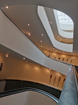 An interior view of a modern architectural structure with multiple curved levels and soft ambient lighting. The design includes overlapping balconies or floors with a smooth, flowing aesthetic. Several ornamental objects resembling large feathers or paddles are mounted on the walls, adding an artistic touch.