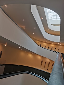 An interior view of a modern architectural structure with multiple curved levels and soft ambient lighting. The design includes overlapping balconies or floors with a smooth, flowing aesthetic. Several ornamental objects resembling large feathers or paddles are mounted on the walls, adding an artistic touch.