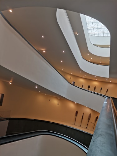 An interior view of a modern architectural structure with multiple curved levels and soft ambient lighting. The design includes overlapping balconies or floors with a smooth, flowing aesthetic. Several ornamental objects resembling large feathers or paddles are mounted on the walls, adding an artistic touch.