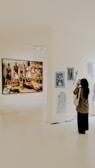 A person is standing in an art gallery, observing an exhibition. A large painting featuring several figures is displayed prominently on a white wall. Nearby, there are smaller black-and-white sketches. The gallery is well-lit, and the floor is polished.