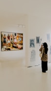 A person is standing in an art gallery, observing an exhibition. A large painting featuring several figures is displayed prominently on a white wall. Nearby, there are smaller black-and-white sketches. The gallery is well-lit, and the floor is polished.