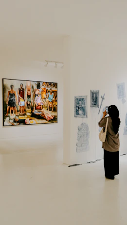 A person is standing in an art gallery, observing an exhibition. A large painting featuring several figures is displayed prominently on a white wall. Nearby, there are smaller black-and-white sketches. The gallery is well-lit, and the floor is polished.