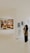 A person is standing in an art gallery, observing an exhibition. A large painting featuring several figures is displayed prominently on a white wall. Nearby, there are smaller black-and-white sketches. The gallery is well-lit, and the floor is polished.