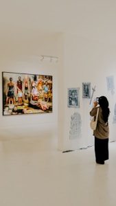 A person is standing in an art gallery, observing an exhibition. A large painting featuring several figures is displayed prominently on a white wall. Nearby, there are smaller black-and-white sketches. The gallery is well-lit, and the floor is polished.