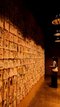 A person is observing a wall covered with numerous pieces of paper or fabric. The papers have colorful drawings and handwritten notes. The room appears dimly lit, creating an intimate and contemplative atmosphere. Industrial-style lights hang from the ceiling, casting a soft glow.