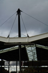 An architectural structure featuring metal and glass elements, with a central black pole supporting cables and beams. The background includes a cloudy sky, creating a contrast with the sleek, modern design of the structure.