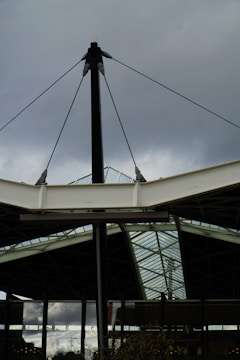 An architectural structure featuring metal and glass elements, with a central black pole supporting cables and beams. The background includes a cloudy sky, creating a contrast with the sleek, modern design of the structure.