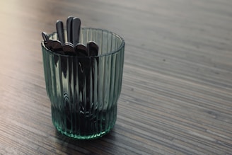 A transparent, ribbed glass cup containing several metal spoons is placed on a wooden surface with a striped pattern. The lighting creates a subtle reflection on the glass and utensils, adding a touch of elegance to the scene.