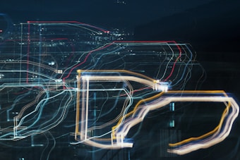 An abstract cityscape with vibrant light trails creating a pattern of lines and shapes against a dark background. The scene suggests movement and energy, as the lights are blurred and layered to form intricate designs.