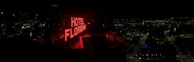 A nighttime cityscape with a prominent red neon sign reading 'HOTEL FLORIDIAN.' The city lights sparkle in the background under a dark sky, adding a lively yet serene ambiance. The architecture is a mix of modern and historical buildings.