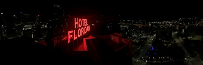 A nighttime cityscape with a prominent red neon sign reading 'HOTEL FLORIDIAN.' The city lights sparkle in the background under a dark sky, adding a lively yet serene ambiance. The architecture is a mix of modern and historical buildings.