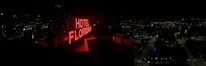 A nighttime cityscape with a prominent red neon sign reading 'HOTEL FLORIDIAN.' The city lights sparkle in the background under a dark sky, adding a lively yet serene ambiance. The architecture is a mix of modern and historical buildings.