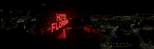 A nighttime cityscape with a prominent red neon sign reading 'HOTEL FLORIDIAN.' The city lights sparkle in the background under a dark sky, adding a lively yet serene ambiance. The architecture is a mix of modern and historical buildings.