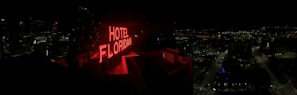 A nighttime cityscape with a prominent red neon sign reading 'HOTEL FLORIDIAN.' The city lights sparkle in the background under a dark sky, adding a lively yet serene ambiance. The architecture is a mix of modern and historical buildings.