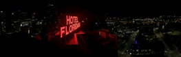 A nighttime cityscape with a prominent red neon sign reading 'HOTEL FLORIDIAN.' The city lights sparkle in the background under a dark sky, adding a lively yet serene ambiance. The architecture is a mix of modern and historical buildings.