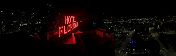 A nighttime cityscape with a prominent red neon sign reading 'HOTEL FLORIDIAN.' The city lights sparkle in the background under a dark sky, adding a lively yet serene ambiance. The architecture is a mix of modern and historical buildings.