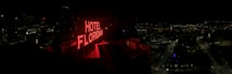 A nighttime cityscape with a prominent red neon sign reading 'HOTEL FLORIDIAN.' The city lights sparkle in the background under a dark sky, adding a lively yet serene ambiance. The architecture is a mix of modern and historical buildings.