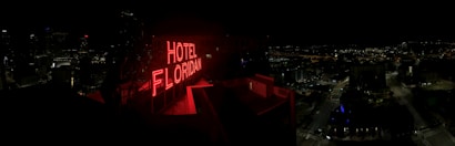 A nighttime cityscape with a prominent red neon sign reading 'HOTEL FLORIDIAN.' The city lights sparkle in the background under a dark sky, adding a lively yet serene ambiance. The architecture is a mix of modern and historical buildings.