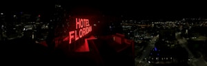 A nighttime cityscape with a prominent red neon sign reading 'HOTEL FLORIDIAN.' The city lights sparkle in the background under a dark sky, adding a lively yet serene ambiance. The architecture is a mix of modern and historical buildings.