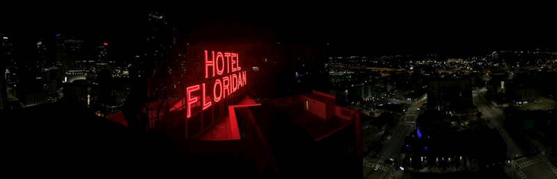 A nighttime cityscape with a prominent red neon sign reading 'HOTEL FLORIDIAN.' The city lights sparkle in the background under a dark sky, adding a lively yet serene ambiance. The architecture is a mix of modern and historical buildings.
