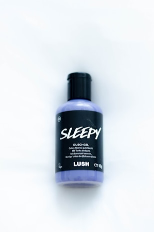 A bottle of Lush Sleepy shower gel is placed on a light surface. The label is primarily black with bold white text, featuring the name 'Sleepy' and product details beneath it. The bottle is a light lavender color and has a black cap.