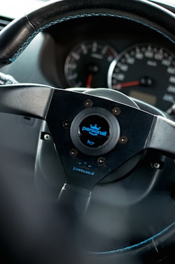 A close-up view of a car steering wheel, prominently featuring a brand logo labeled 'personal' in the center. The dashboard in the background shows the speedometer and other gauges.