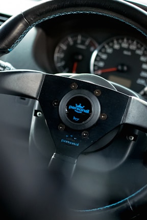 A close-up view of a car steering wheel, prominently featuring a brand logo labeled 'personal' in the center. The dashboard in the background shows the speedometer and other gauges.