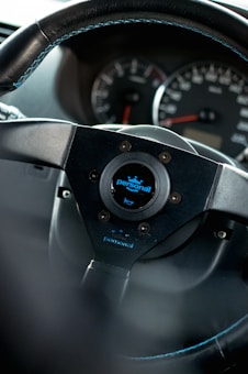 A close-up view of a car steering wheel, prominently featuring a brand logo labeled 'personal' in the center. The dashboard in the background shows the speedometer and other gauges.