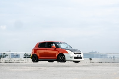 Red compact car parked near a park with city skyline.