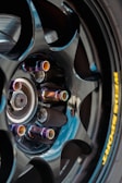 A close-up view of a glossy black alloy wheel with a multi-spoke design. The lug nuts have an iridescent finish, creating a colorful effect. A portion of a brake disc is visible behind the wheel. The tire displays a yellow brand logo imprinted on the sidewall.