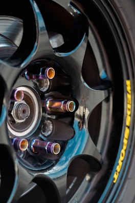 A close-up view of a glossy black alloy wheel with a multi-spoke design. The lug nuts have an iridescent finish, creating a colorful effect. A portion of a brake disc is visible behind the wheel. The tire displays a yellow brand logo imprinted on the sidewall.