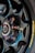 A close-up view of a glossy black alloy wheel with a multi-spoke design. The lug nuts have an iridescent finish, creating a colorful effect. A portion of a brake disc is visible behind the wheel. The tire displays a yellow brand logo imprinted on the sidewall.