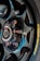 A close-up view of a glossy black alloy wheel with a multi-spoke design. The lug nuts have an iridescent finish, creating a colorful effect. A portion of a brake disc is visible behind the wheel. The tire displays a yellow brand logo imprinted on the sidewall.