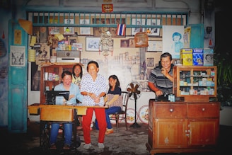 A lively scene with several people enjoying themselves in a retro-style shop or home setting. There are various household items around, including a sewing machine and merchandise displayed in wooden cabinets. The walls are adorned with vintage posters and photographs, and the atmosphere appears to be cheerful and nostalgic.