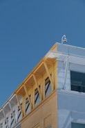 A modern building facade with a combination of light blue, yellow, and white colors. Metal structural elements support glass windows, and a security camera is mounted at the top corner. Shadows and reflections are visible on the building surfaces.