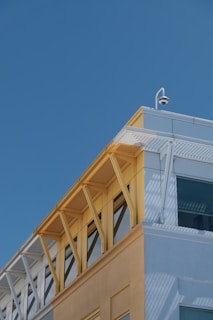 A modern building facade with a combination of light blue, yellow, and white colors. Metal structural elements support glass windows, and a security camera is mounted at the top corner. Shadows and reflections are visible on the building surfaces.