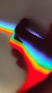 A hand is positioned with a spectrum of rainbow colors projected across it, creating a vibrant, colorful effect against a plain background. The colors transition smoothly from red to violet, highlighting the contours of the hand in silhouette.